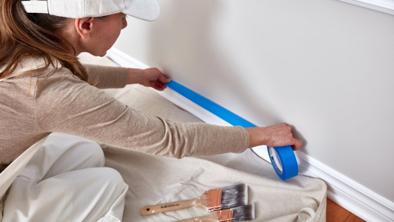 Woman taping wall to paint trim (Photo by kurhan / Shutterstock.com)