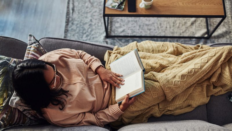 A woman wrapped in blanket reading a book on the sofa at home (Photo by LumiNola/E+ via Getty Images)