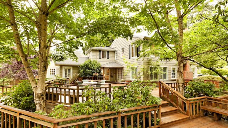 a large brown deck interspersed with bushes and trees in the backyard of a large white house