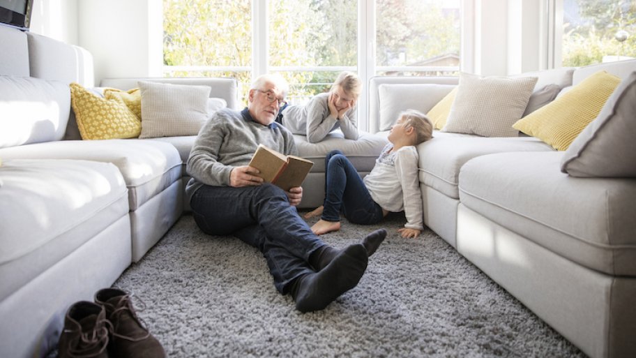 elderly man and two young kids read book in front of large windows between two white couches