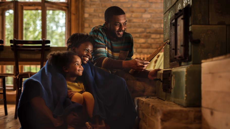 woman, man, and child in cozy blankets huddled next to furnace in low light and smiling