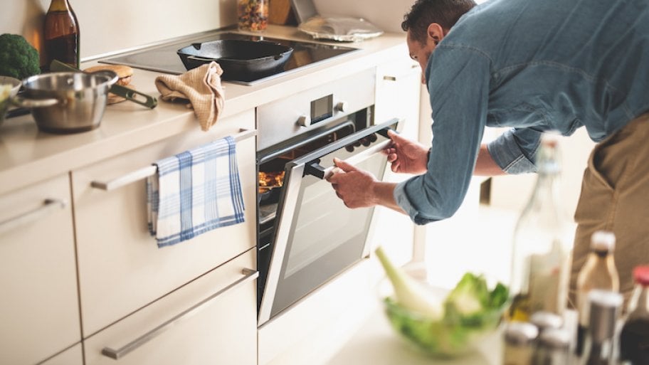man opening oven to check on food inside in bright, cream colored kitchen strewn with various cooking tools