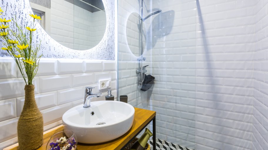Modern small bathroom with white tile walls, glass rain shower, backlit round mirror, and vessel sink on narrow wood table