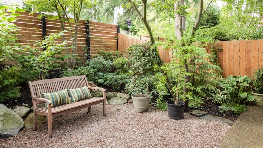 a light wood privacy fence surrounds a private yard with plants, bushes, a bench, and gravel