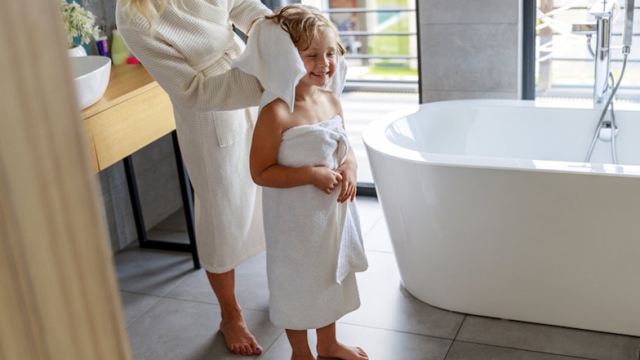 child in white towel gets hair dried by adult in bathroom with gray tiled floors and white bathtub
