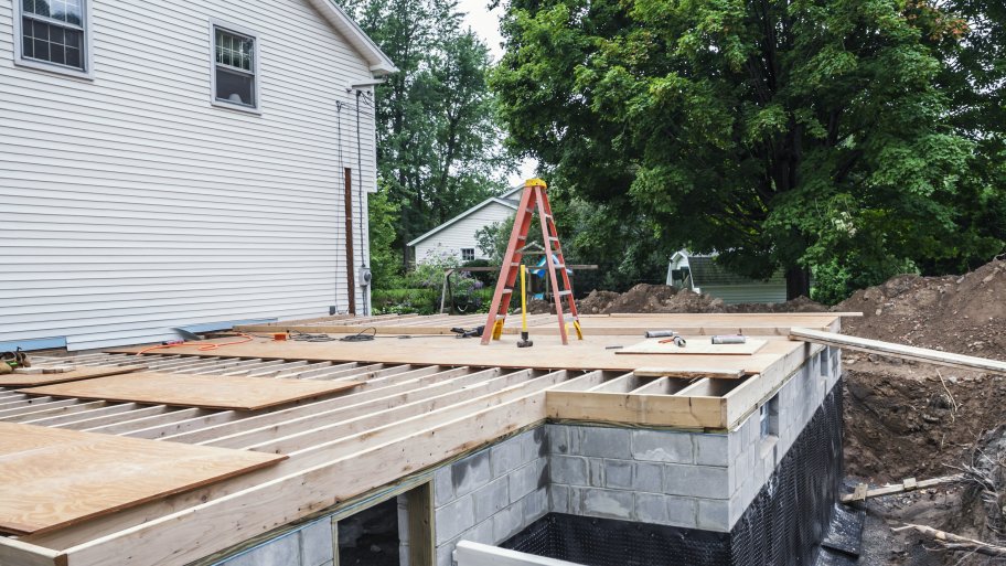 The subfloor covering a new home addition basement foundation is in process of being built at a suburban residential in-law apartment construction building site. Various tools, construction material and a ladder are strewn about.