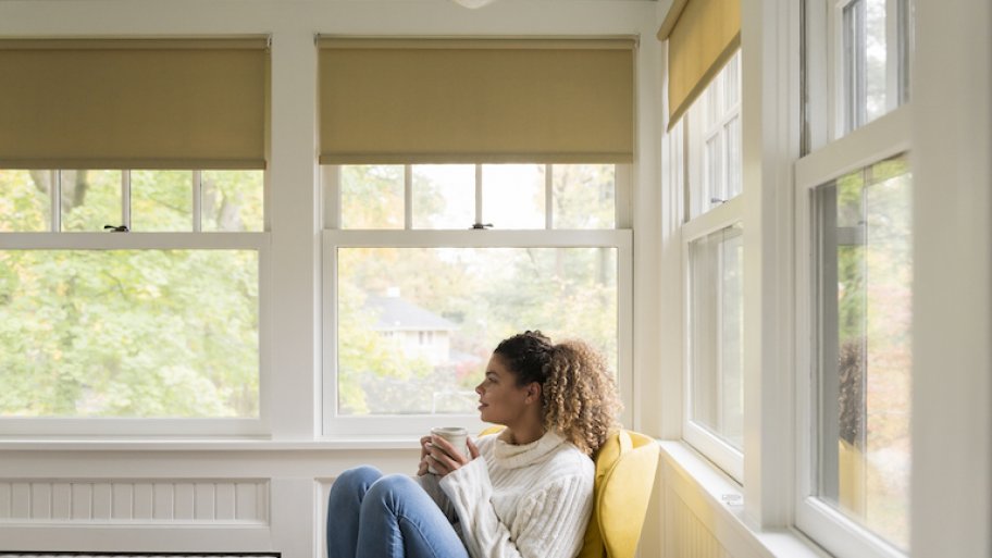 woman sits in corner of room filled with double hung windows holding cup of coffee and lots of greenery outside