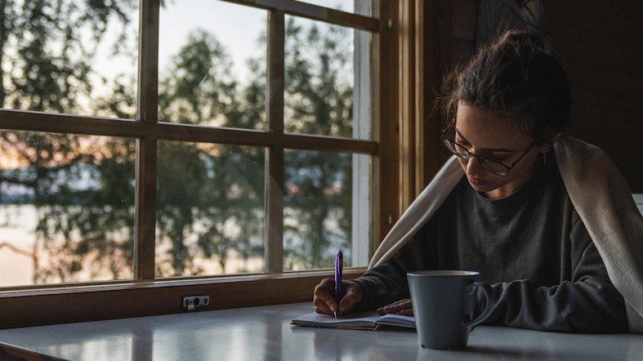 woman with pen, paper, and cup of coffee sits next to window in early morning