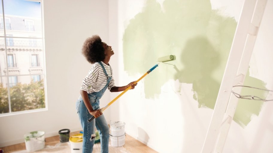 a woman in overalls painting a white wall with green paint and smiling