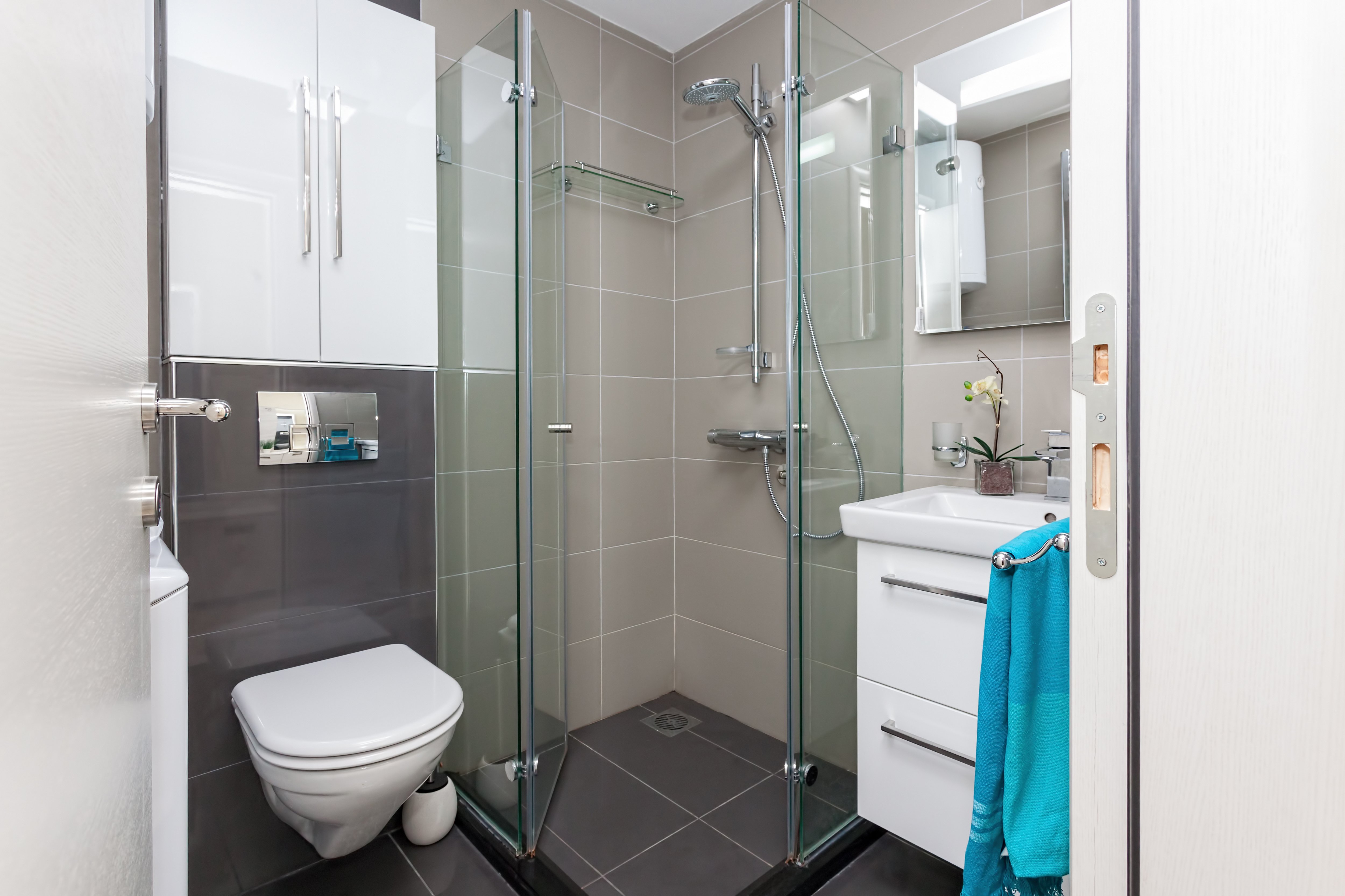 Small modern bathroom with gray tiles, glass shower, wall-hung toilet, and white vanity and cabinets