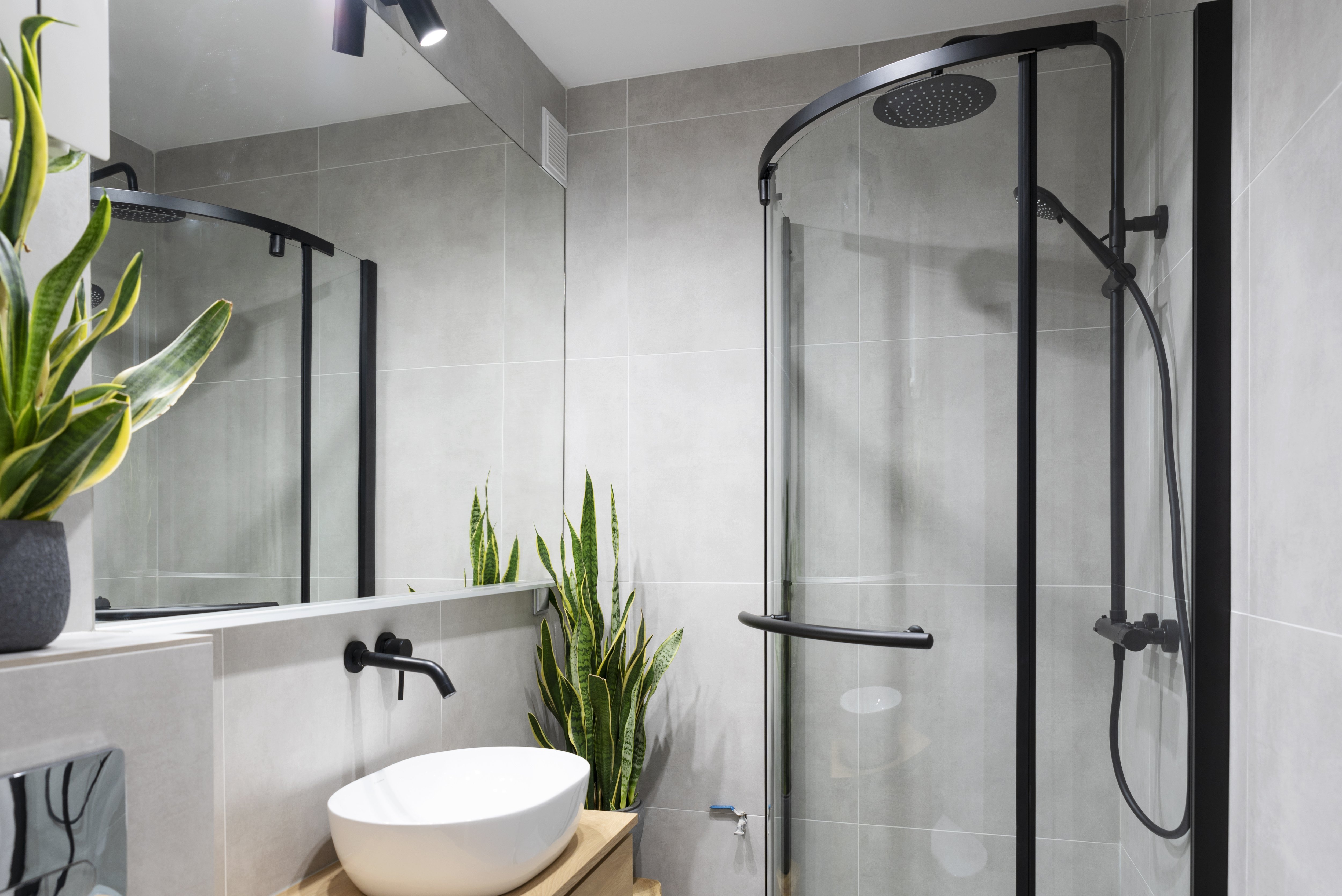 Small bathroom with gray tiles, glass and black metal shower, wall-mounted faucet, vessel sink, and plants
