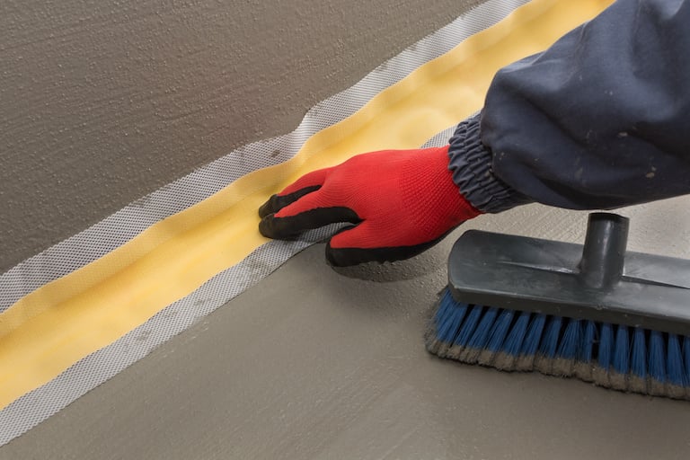 a hand with red gloves applies waterproofing tape to bottom corner of room