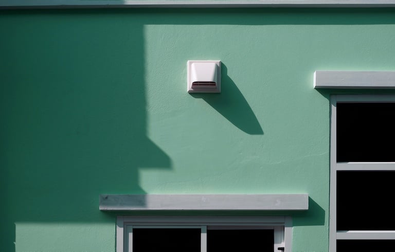 White dryer vent and part of window frames with sunlight and shadow on surface of pale green cement wall background of house building