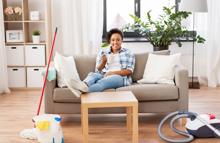 woman relaxes on couch with cleaning supplies on the floor next to her. She smiles and holds a coffee cup.