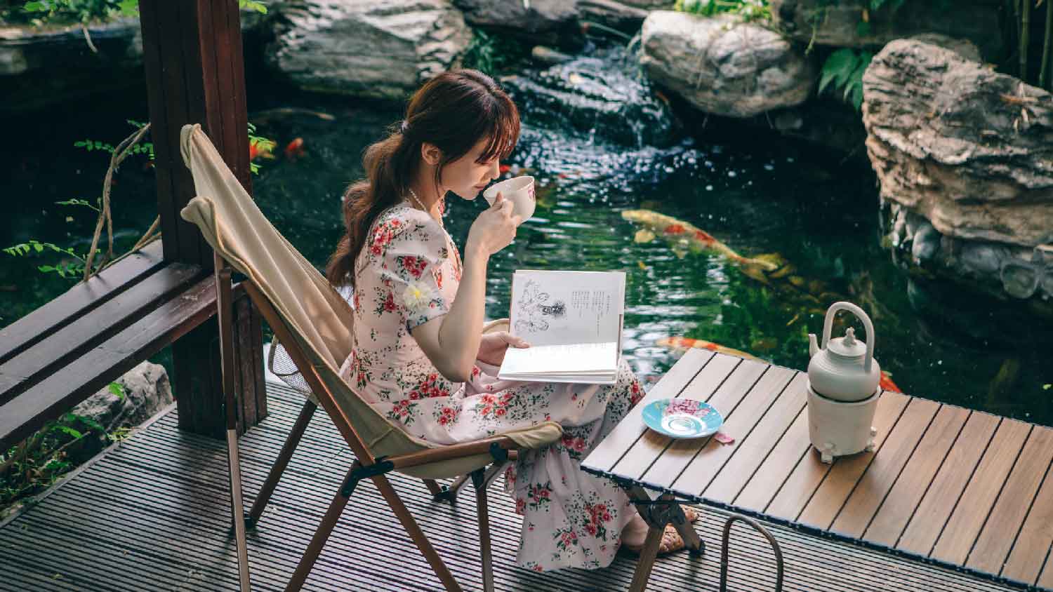 A young woman reading a book under a pergola with a koi pond next to her