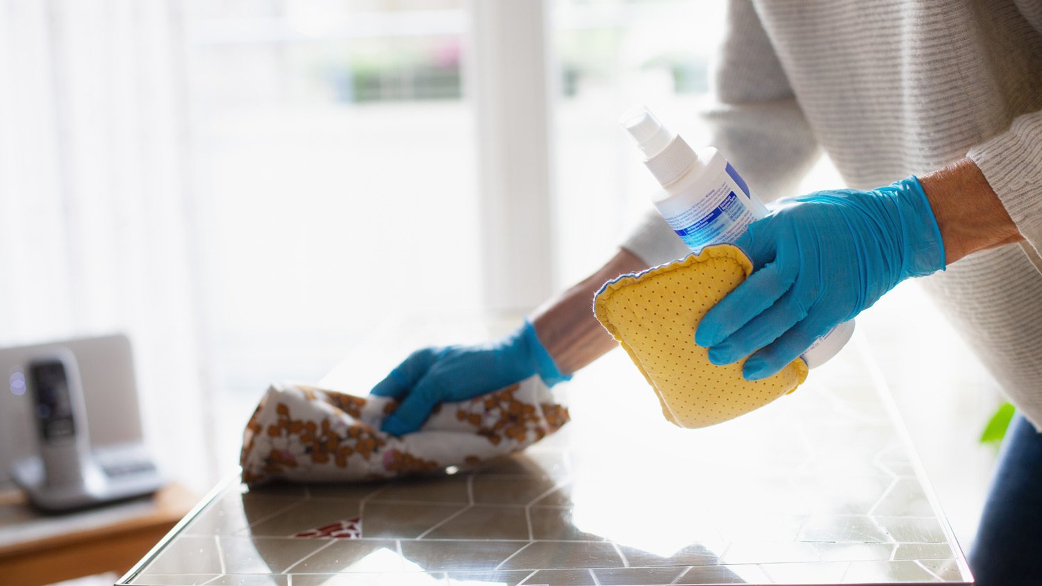 A woman’s hands in gloves cleaning a table with disinfectant spray