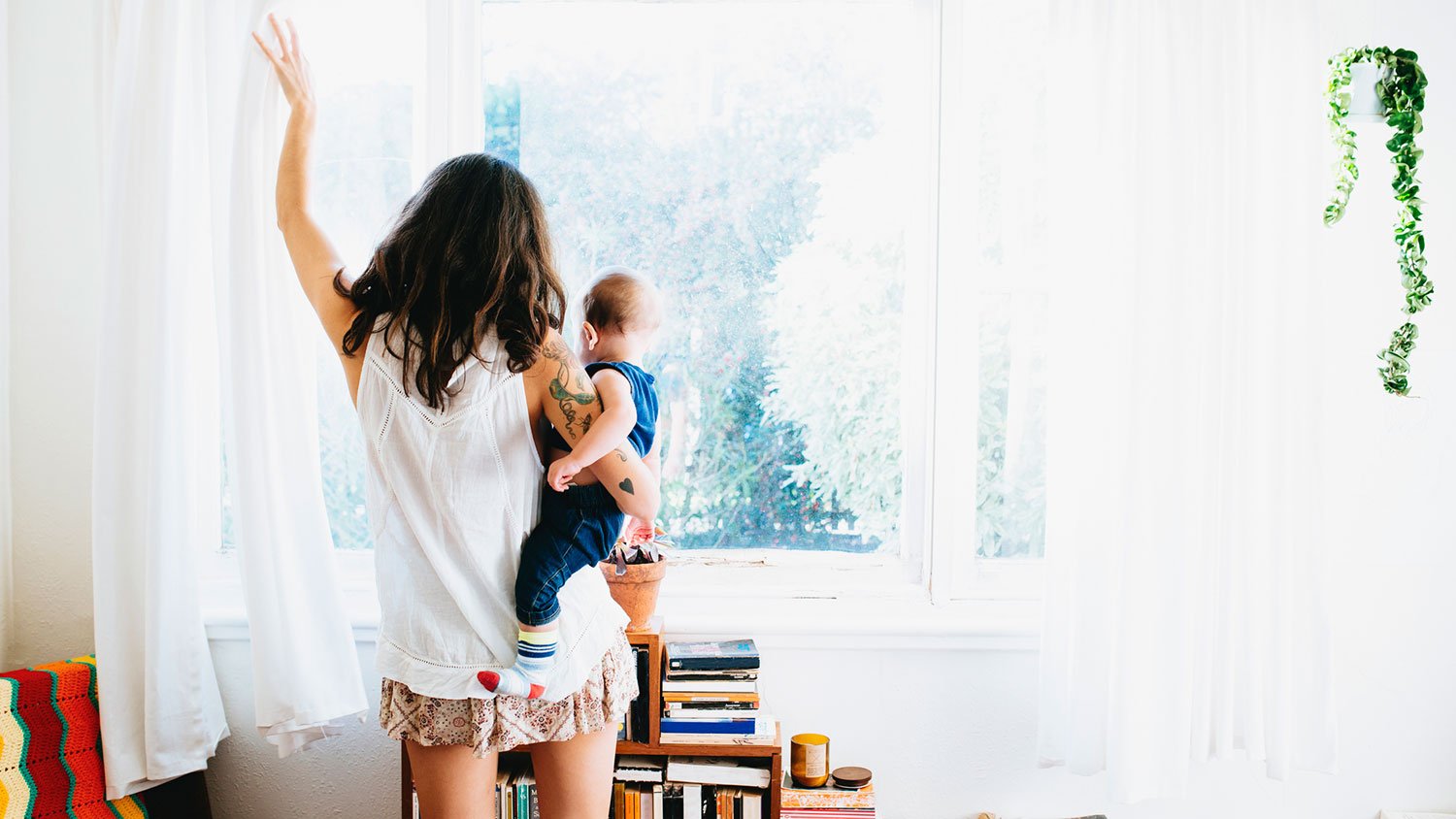 Woman holding a baby is opening the living room curtains 