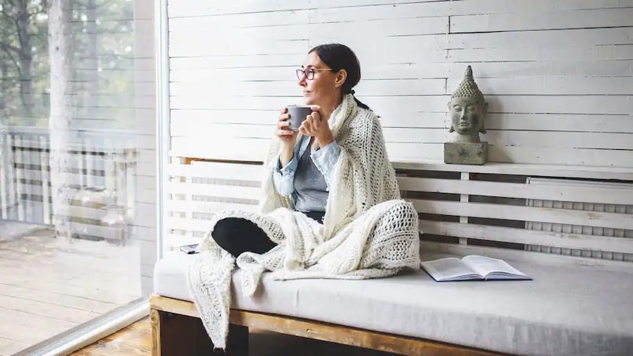 Woman sitting by the window wrapped in a blanket drinking coffee