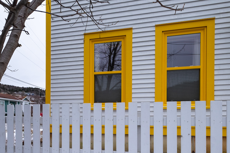 two double hung windows with yellow frames on a white house with a white picket fence