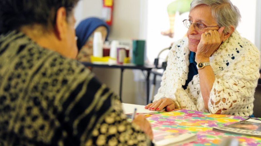 elderly woman sits at a table with a man who is writing.